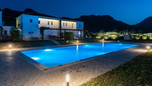 Una piscina con borde de piedra, un edificio con múltiples ventanas y un balcón, y montañas al fondo.