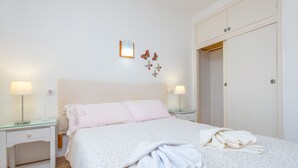 A neatly made bed with a white and light pink bedspread, two bedside tables with lamps, a wall-mounted light fixture, and a wardrobe with a door. - SOL - Nice apartment close to the beach and leisure area. Free WiFi. (Alcúdia)