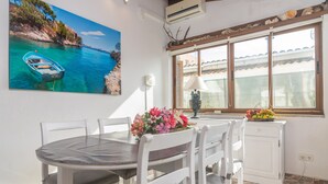Uma sala de jantar com uma mesa redonda, cadeiras brancas, um arranjo de flores e uma janela com vista para a paisagem costeira.