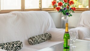 Um quarto com um sofá branco, uma almofada estampada, um vaso com flores, uma garrafa de champanhe e duas taças sobre uma mesa.