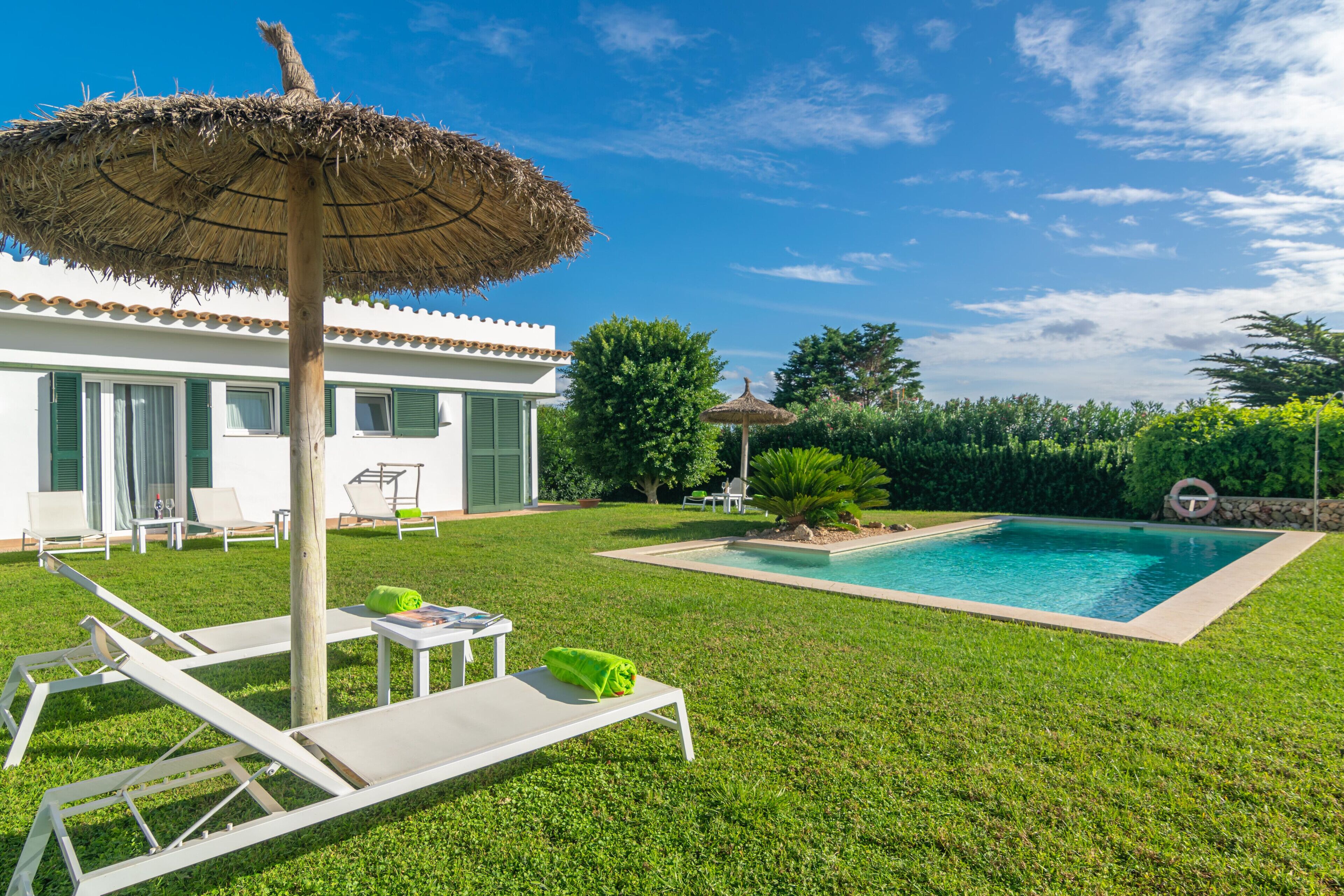 Un área de piscina con sillones de playa y una sombrilla de paja, rodeada por un césped bien cuidado y un edificio blanco con contraventanas verdes.