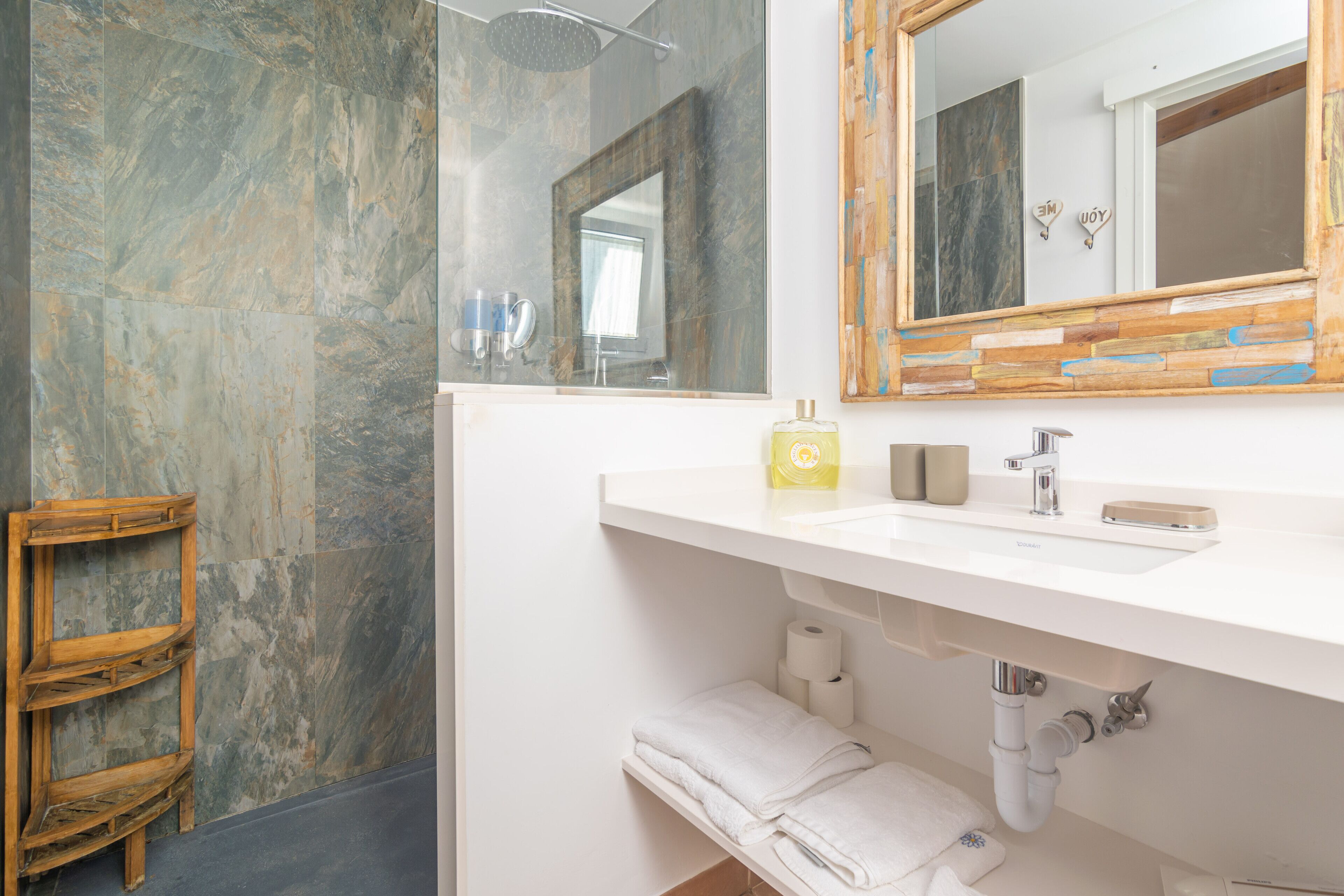 Un baño moderno con pared de mármol, estante de madera, lavamanos blanco y espejo con marco de madera.