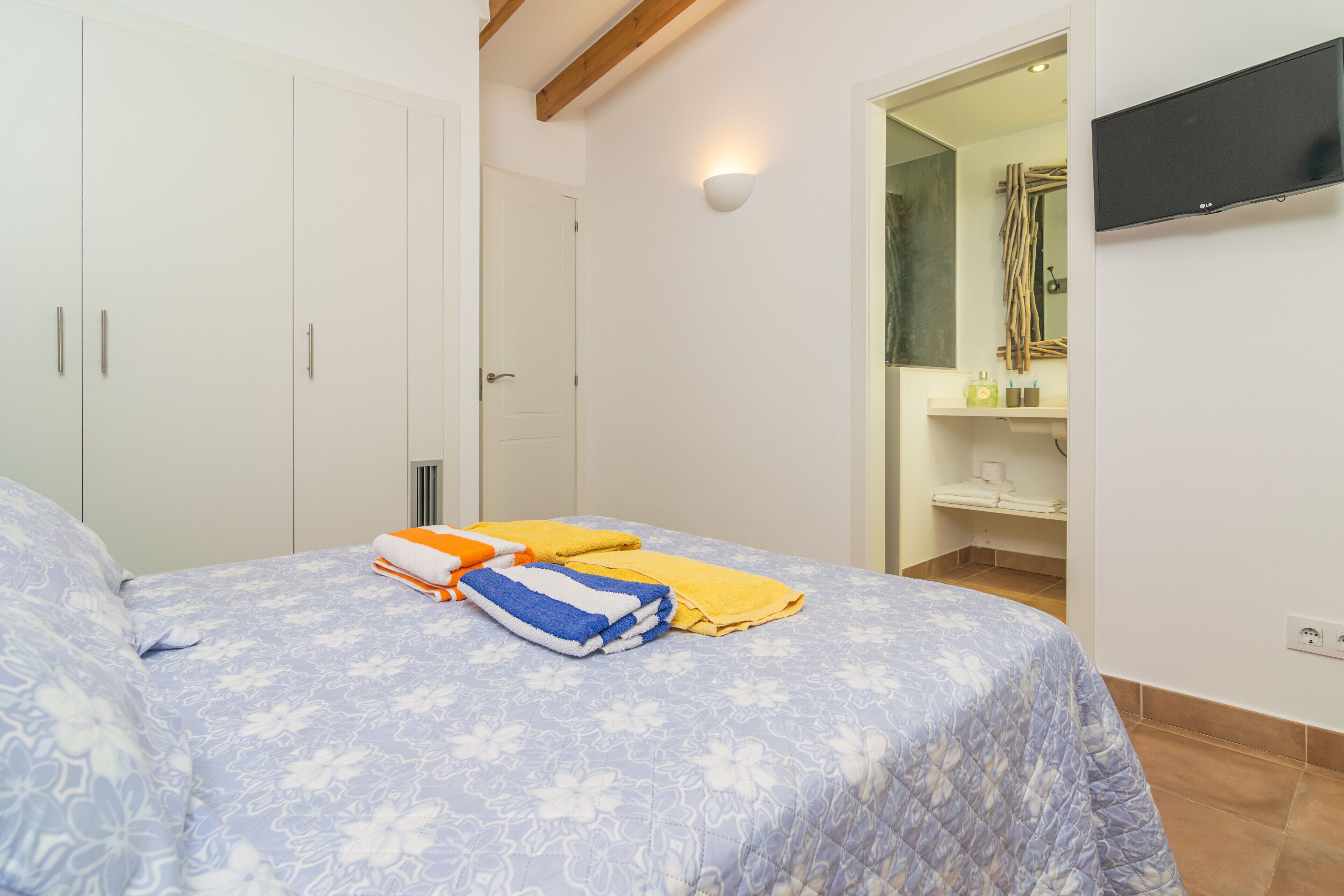 Un dormitorio con cama, armario, televisor y baño visible a través de una puerta abierta.