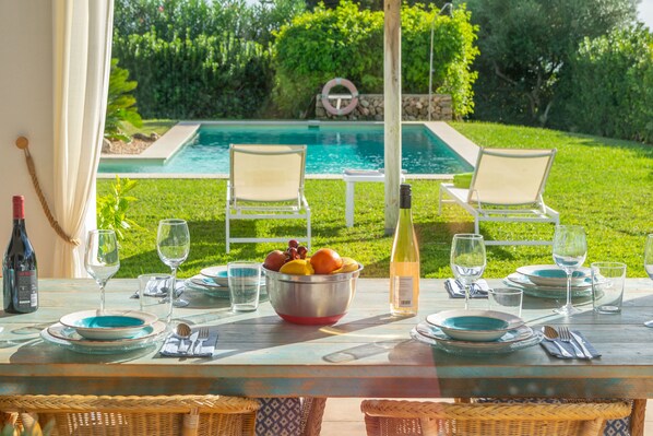 An outdoor dining area with a pool view, a table set with plates, glasses, and a bowl of fruit.