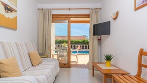 A hotel room with a bed, a TV, a small table with a plant, and a view of a swimming pool and greenery outside. - VILLA VICTORIA - Charming house with shared pool. (Alaior)