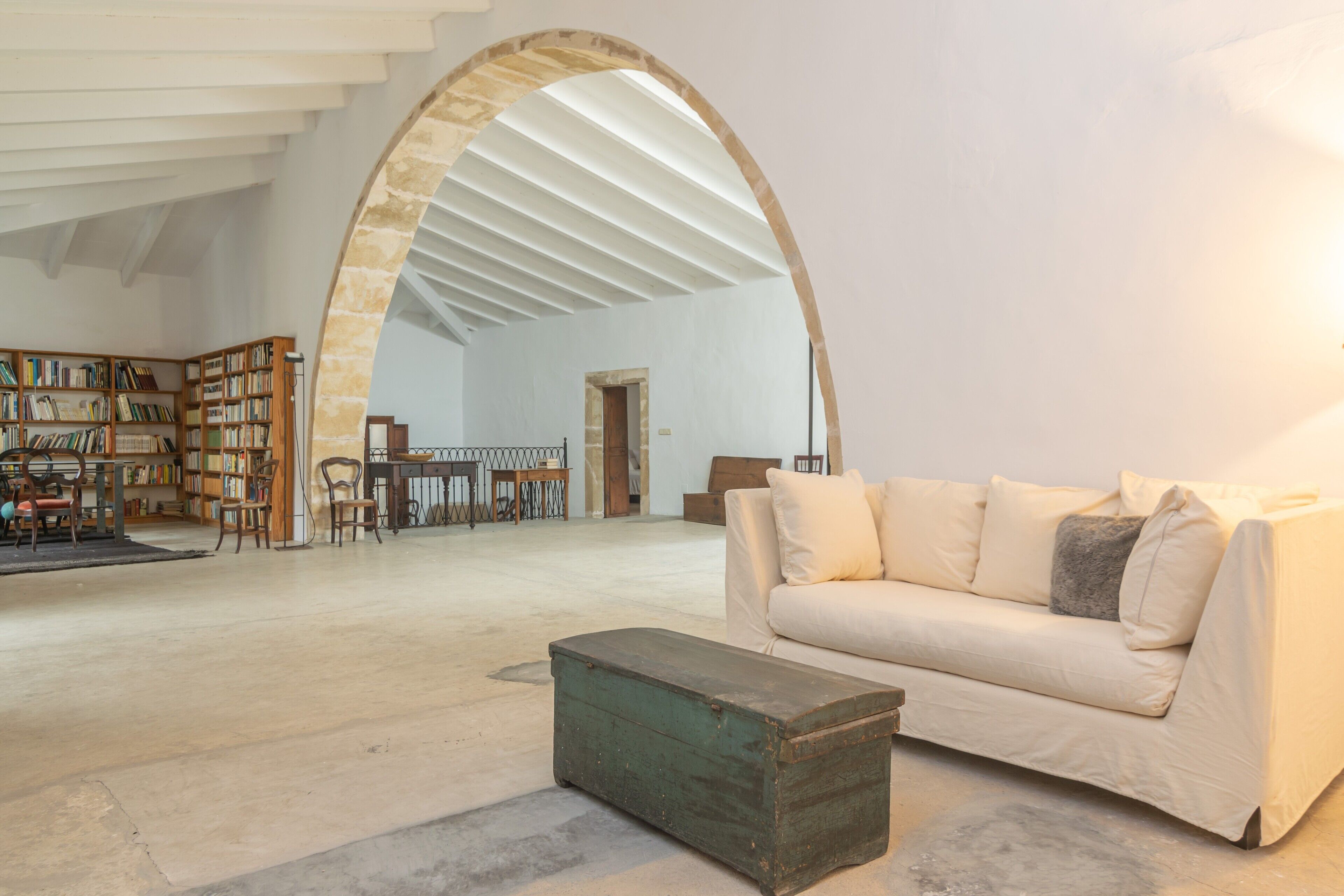 Une pièce spacieuse avec un plafond haut, une grande porte en arc de cercle, un canapé blanc, une commode en bois et une bibliothèque remplie de livres.