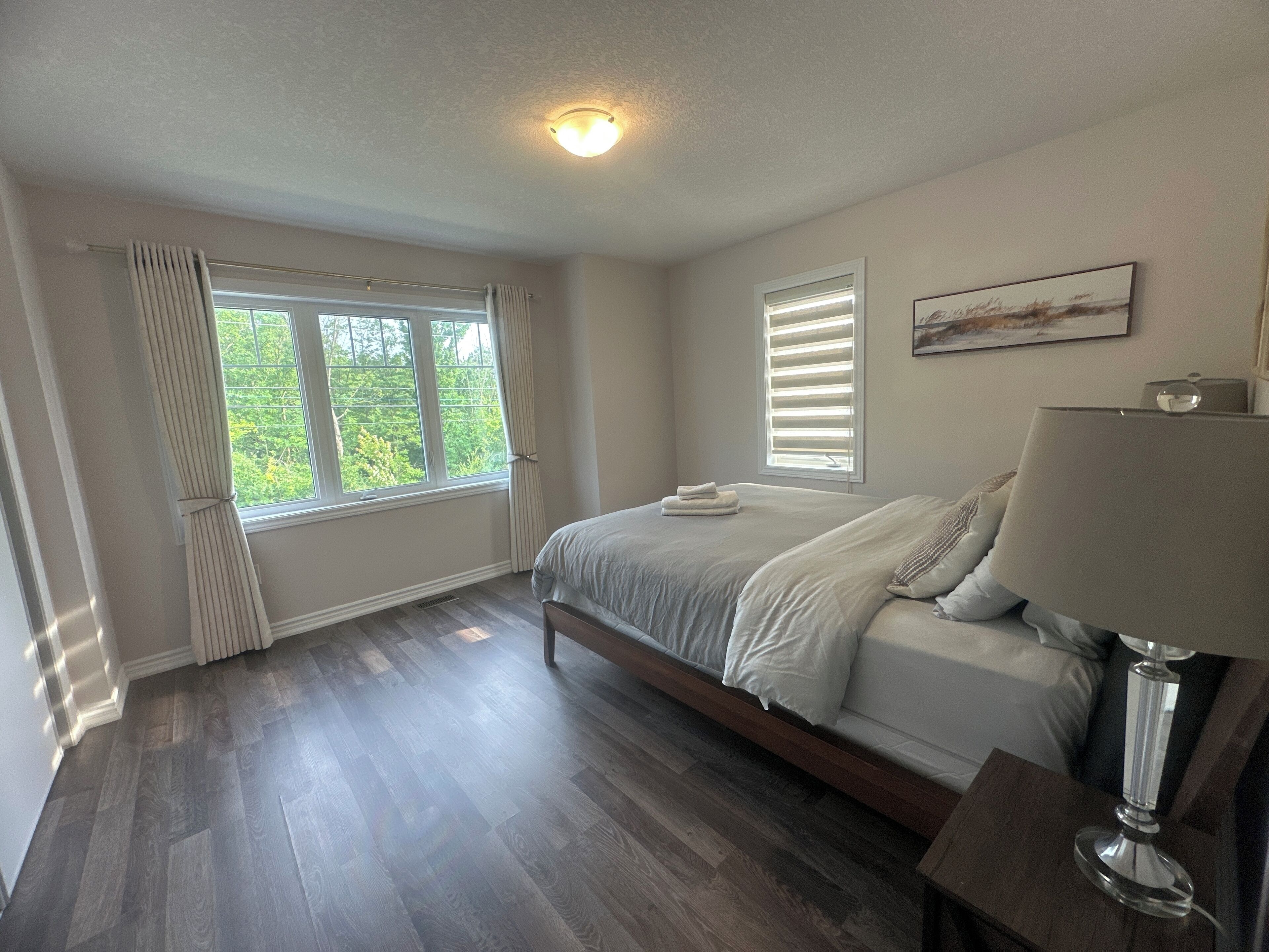 A bedroom with a bed, a bedside table, a lamp, a window with blinds, and a ceiling light.