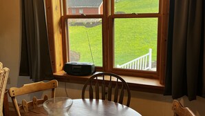 Un comedor con una mesa y sillas de madera, una vela y vistas a un jardín de césped y un edificio rojo a través de un ventanal con marcos de madera.