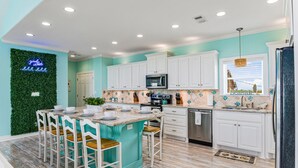 A kitchen with teal walls, white cabinets, a central island with bar stools, a neon sign, and a view through the window. - Sweetheart savings! 3 min walk to Beach, Ocean View, WIFI, King 🛏 (Crystal Beach)