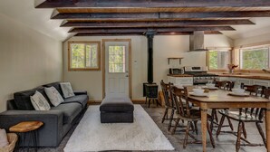 Un amplio salón con un sofá gris, un tapete blanco y una mesa de comedor con sillas de madera. Se ve una cocina con estufa y microondas al fondo.