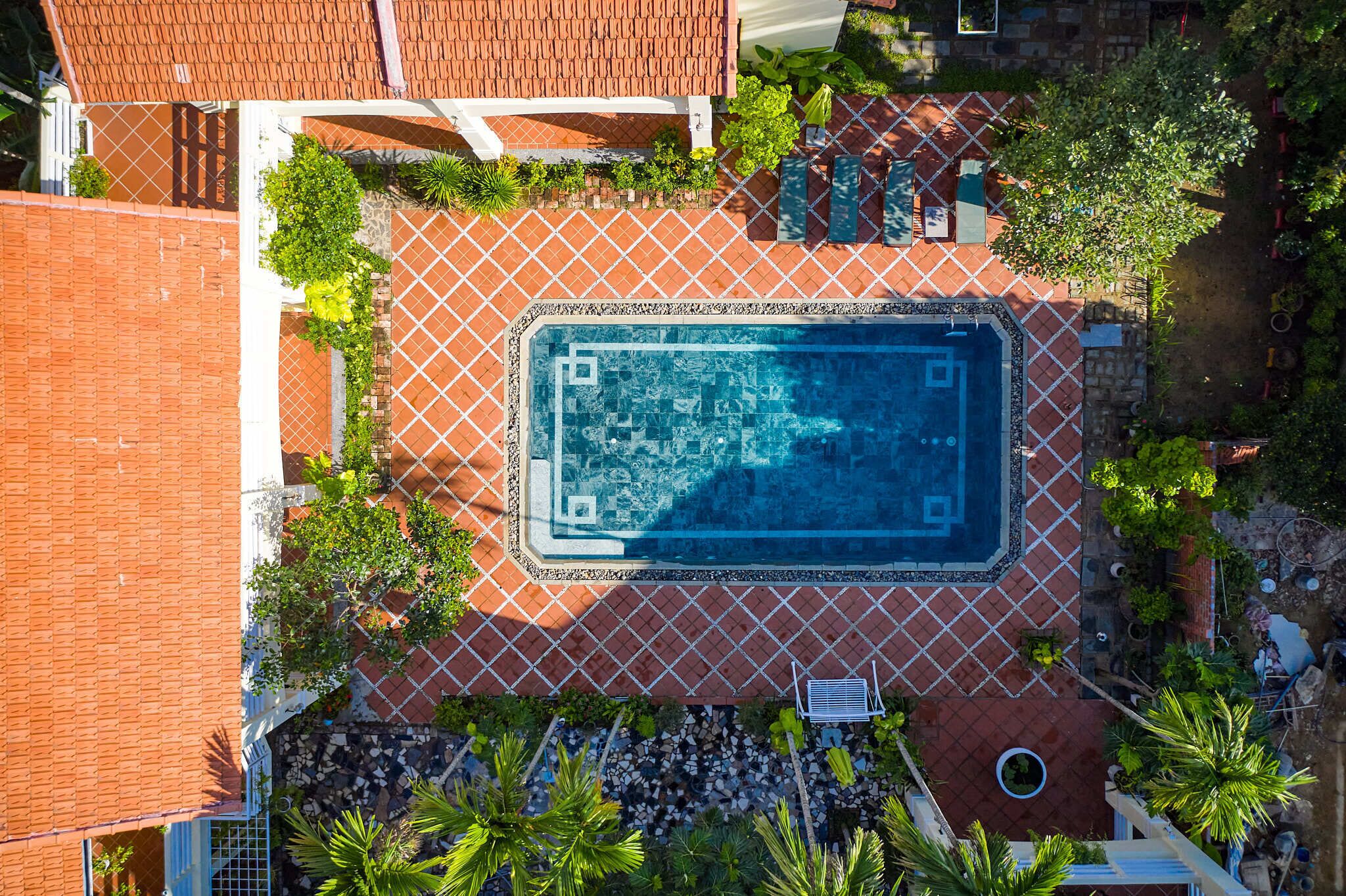 Vista aerea di una piscina circondata da un patio piastrellato, con recinzione e vegetazione.