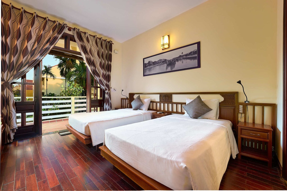Habitación de hotel con dos camas, un balcón con cortinas, un cuadro en la pared y una mesita de noche de madera.