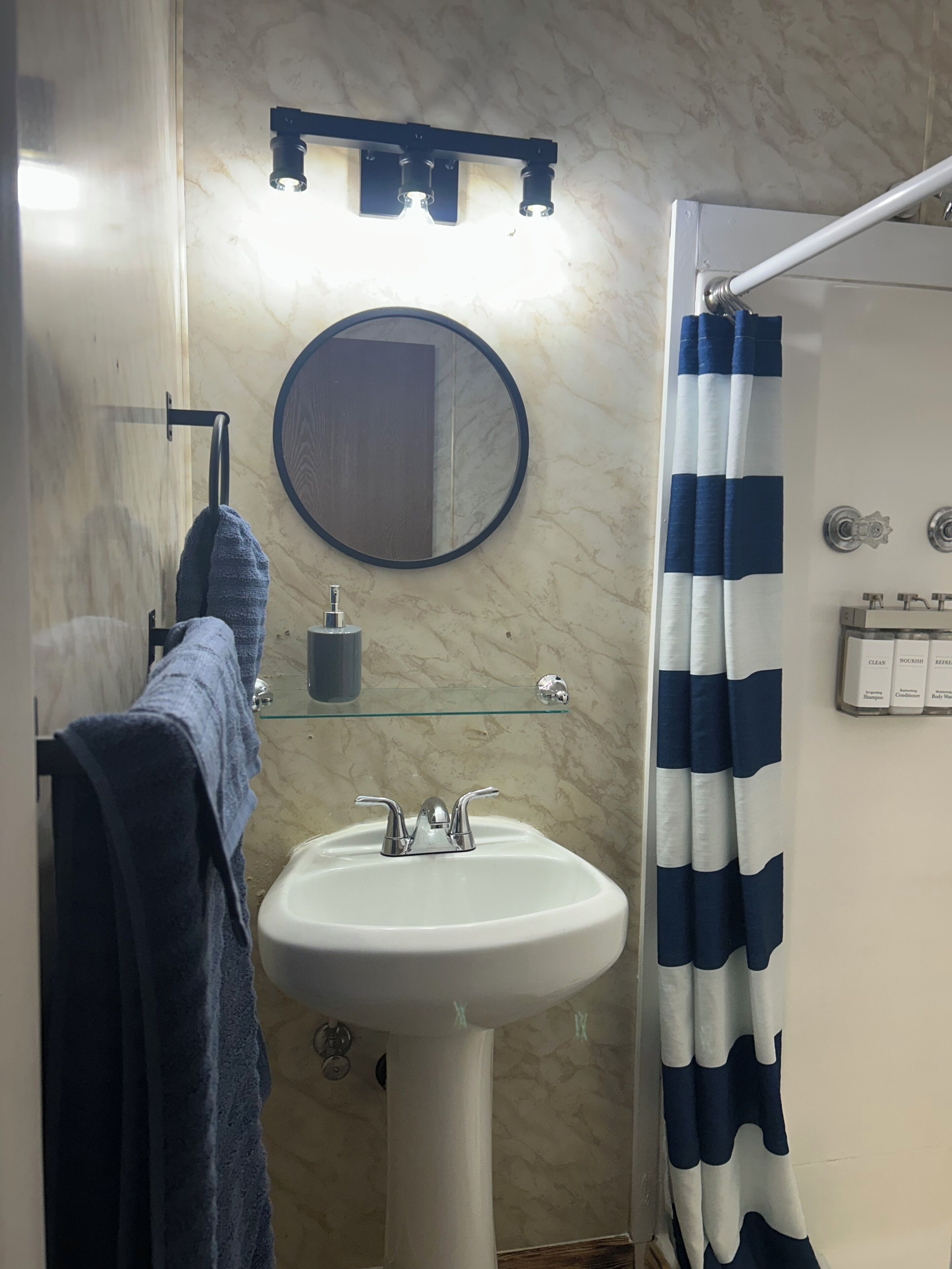 Une salle de bain équipée d’un lavabo, d’un miroir, d’un porte-serviettes, d’un rideau de douche et d’une étagère en verre où sont rangés les articles de toilette.