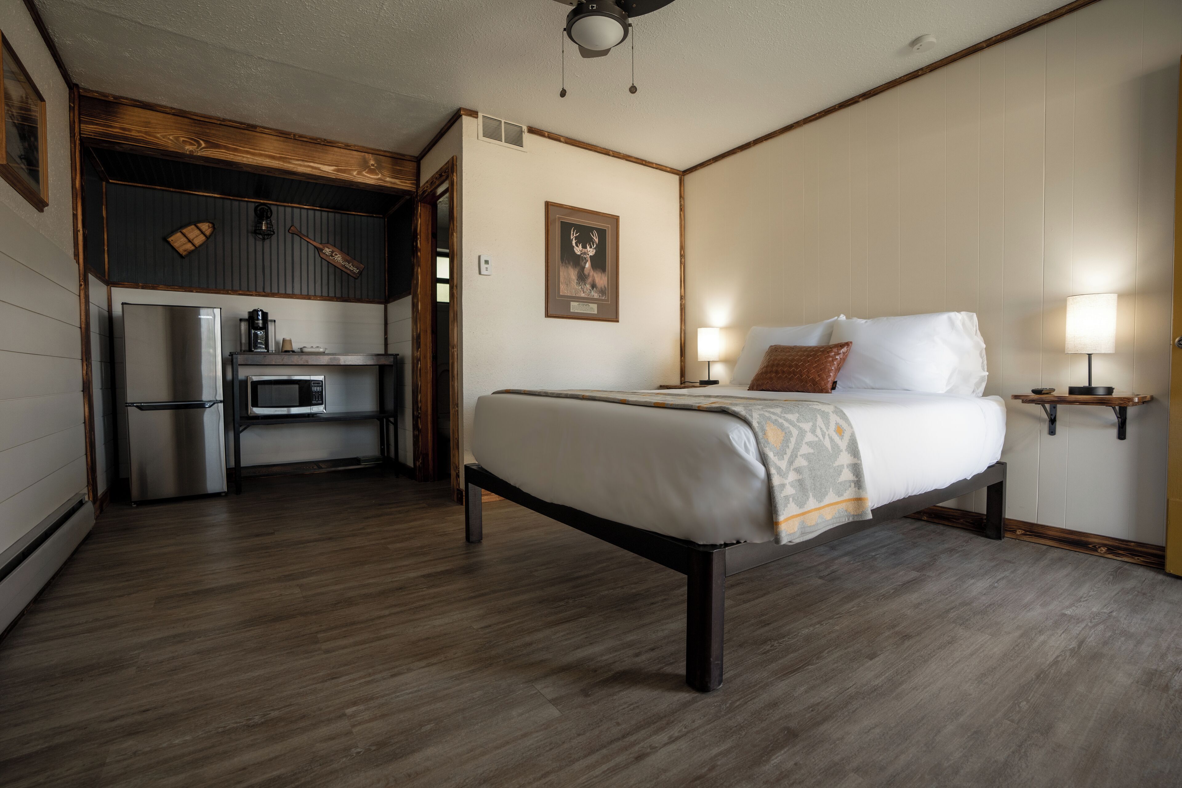 A hotel room with a bed, bedside tables, a desk with a chair, a refrigerator, a microwave, and a ceiling fan.