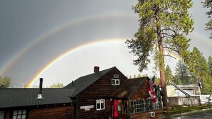 A wooden cabin with a chimney, a porch with outdoor seating, and a rainbow in the background. - Columbia Point Resort (Kettle Falls)