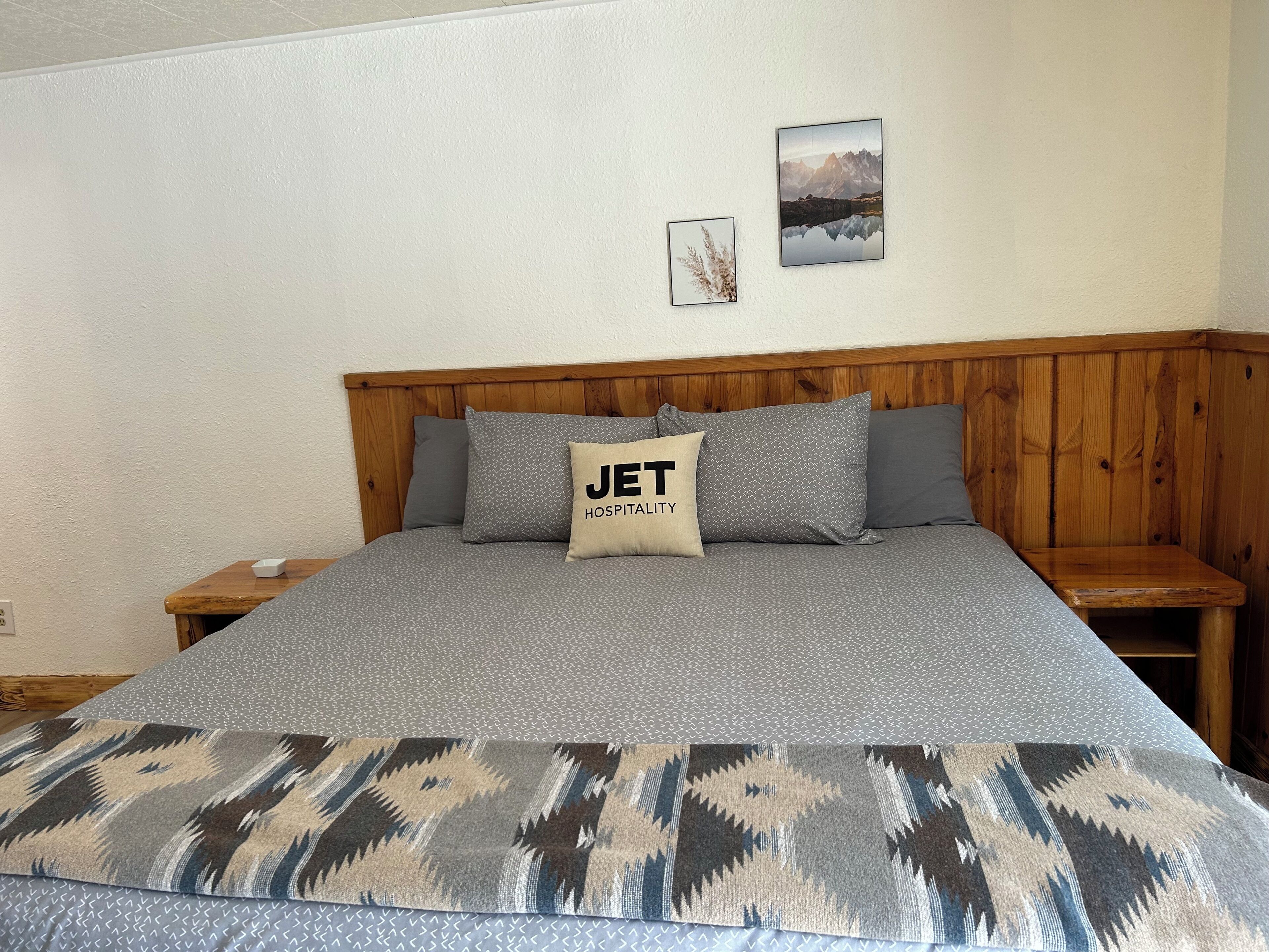A bed with a grey patterned bedspread, a pillow with 'JET HOSPITALITY' text, a wooden headboard, and two framed pictures on the wall.