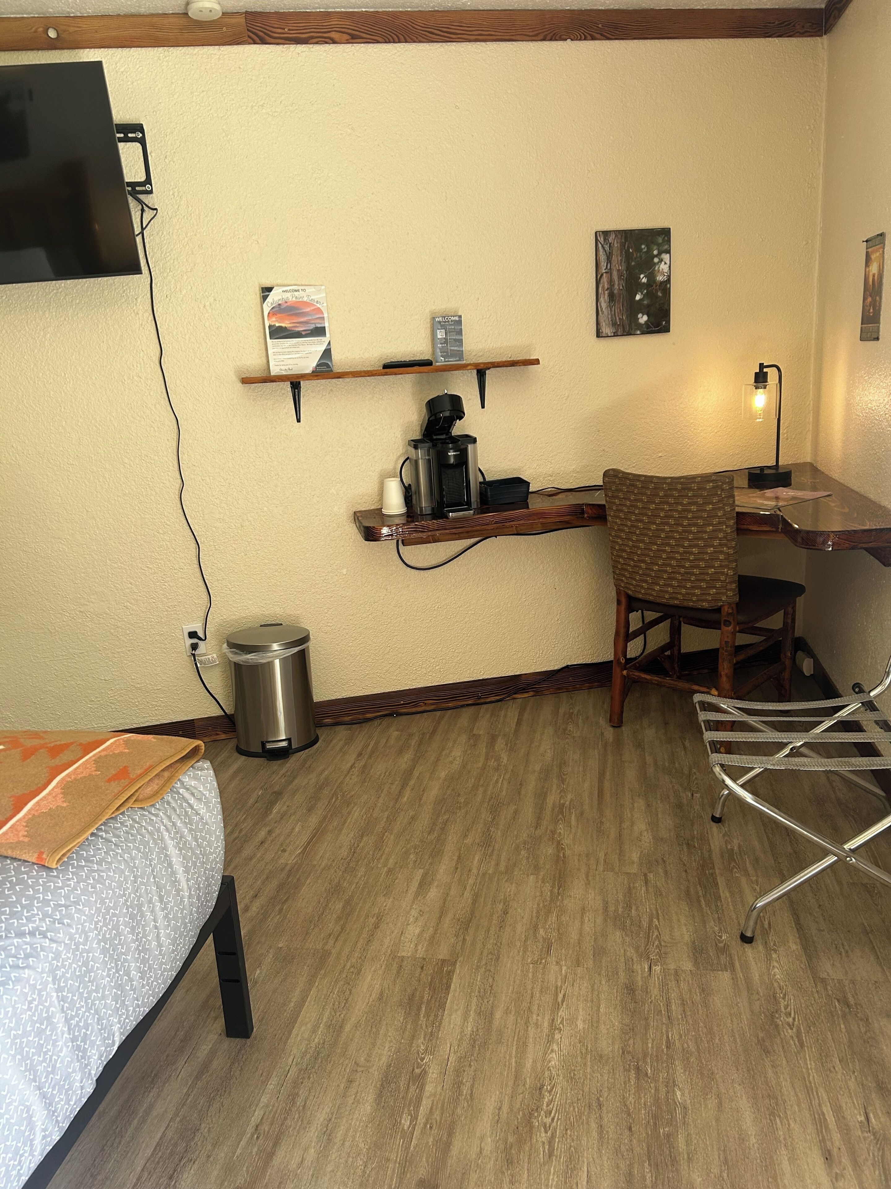 Une petite pièce agréable et confortable, avec un lit, un bureau doté d’une machine à café, une chaise, une lampe et un téléviseur fixé au mur.