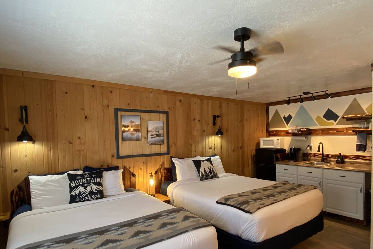 A hotel room with two beds, a ceiling fan, wooden walls, a kitchen area, and a framed picture on the wall.