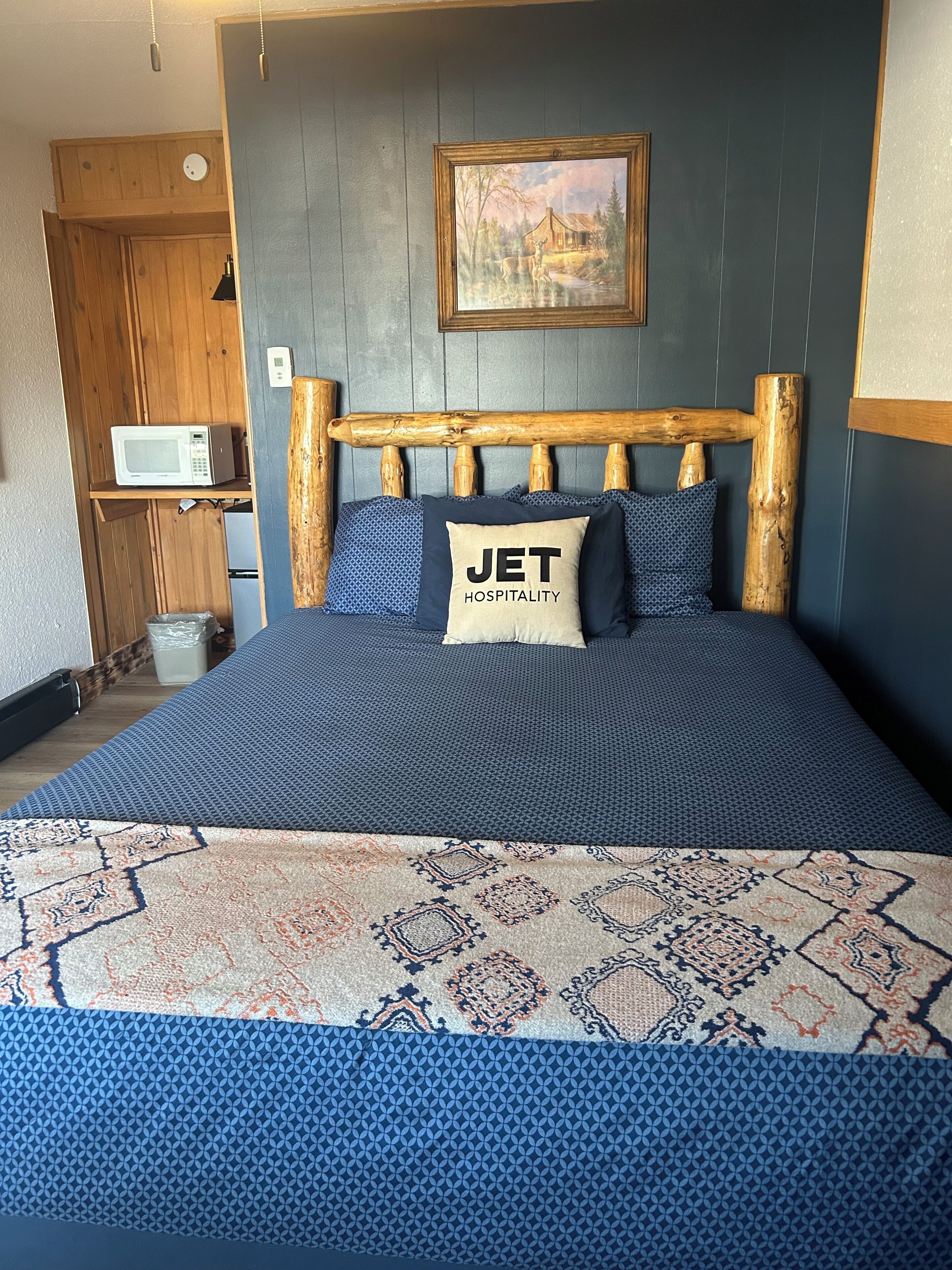 A bed with a wooden headboard, a painting above it, and a pillow with 'JET HOSPITALITY' text.