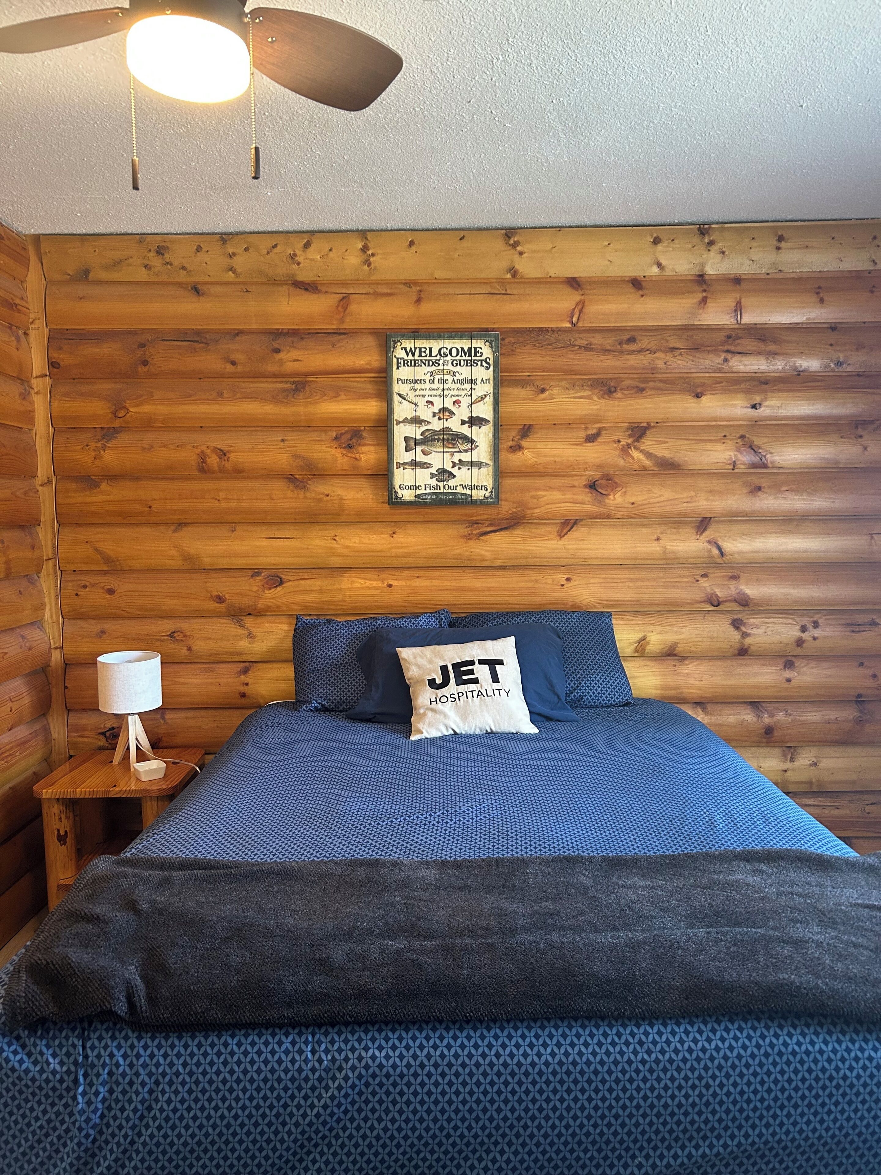 Ein holzverkleideter Raum im Chalet-Stil mit einem Bett, einem Wandzeichen, einem Deckenventilator und einem kleinen Nachttisch mit Lampe.