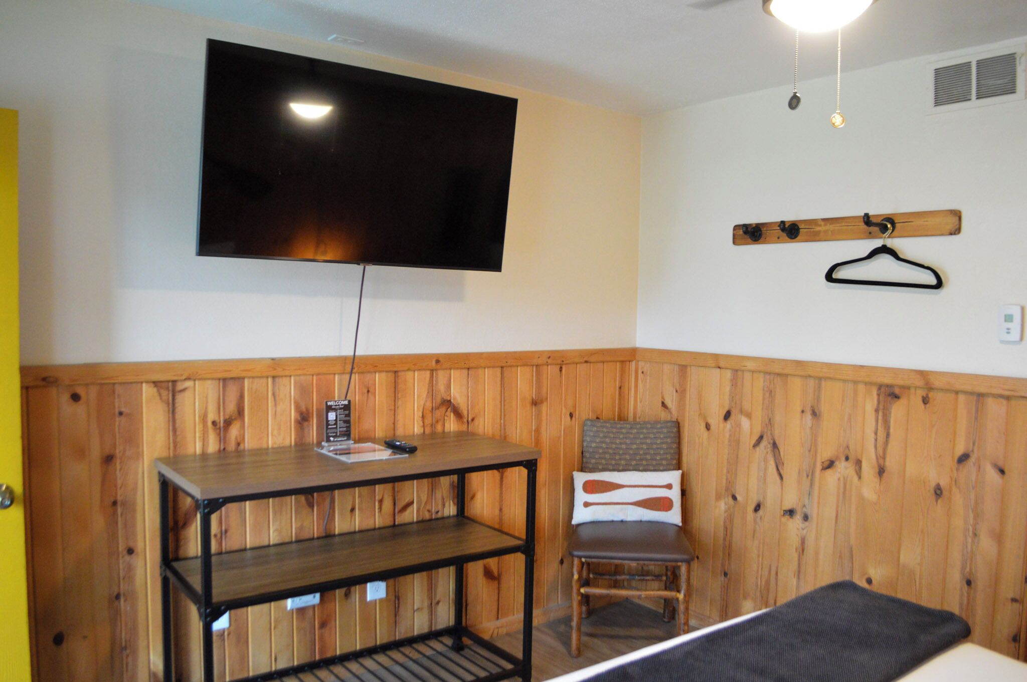 A room with a wooden paneled wall, a mounted TV, a wooden table with a remote control, a chair with a patterned cushion, and a coat rack with a hanger.