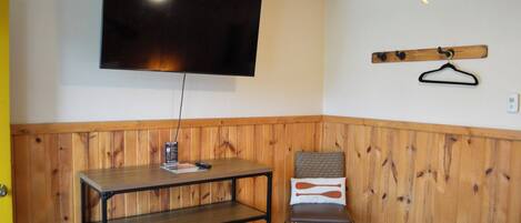 A room with a wooden paneled wall, a mounted TV, a wooden table with a remote control, a chair with a patterned cushion, and a coat rack with a hanger.