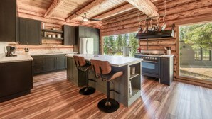 A modern kitchen with wooden beams, a large island with bar stools, a built-in stove, and a view of the outdoors. - Tahoe Wanderer - Super Cool Luxury Log Cabin with NEW Kitchen, Private Hot Tub (Tahoe Vista)