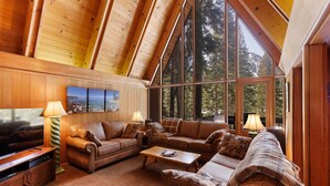 A cozy living room with wooden walls and a sloped ceiling, featuring a large window with a view of trees and a fireplace. - Snowy Paws Alpine Meadows - Ski Shuttle- Dog OK - Close to Slopes - Amazing Views (Olympic Valley)