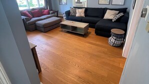 Un salón con un sofá de color oscuro, una mesa de centro de madera y una mesita redonda. Hay cuadros enmarcados en la pared y una ventana con cortinas.