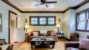 A cozy living room with a brown leather sofa, a coffee table, and a ceiling fan. There are two windows with curtains, a door, and a small side table with a lamp.