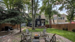A backyard patio with a fire pit and Adirondack chairs, surrounded by a brick patio and a wooden fence.