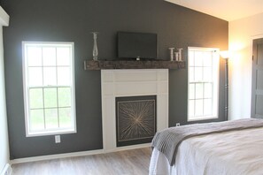 A bedroom with a fireplace, two windows, a television, and a lamp.