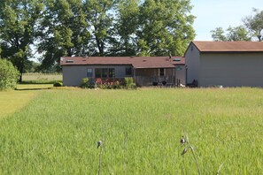 A single-story house with a porch, surrounded by a grassy field and trees.