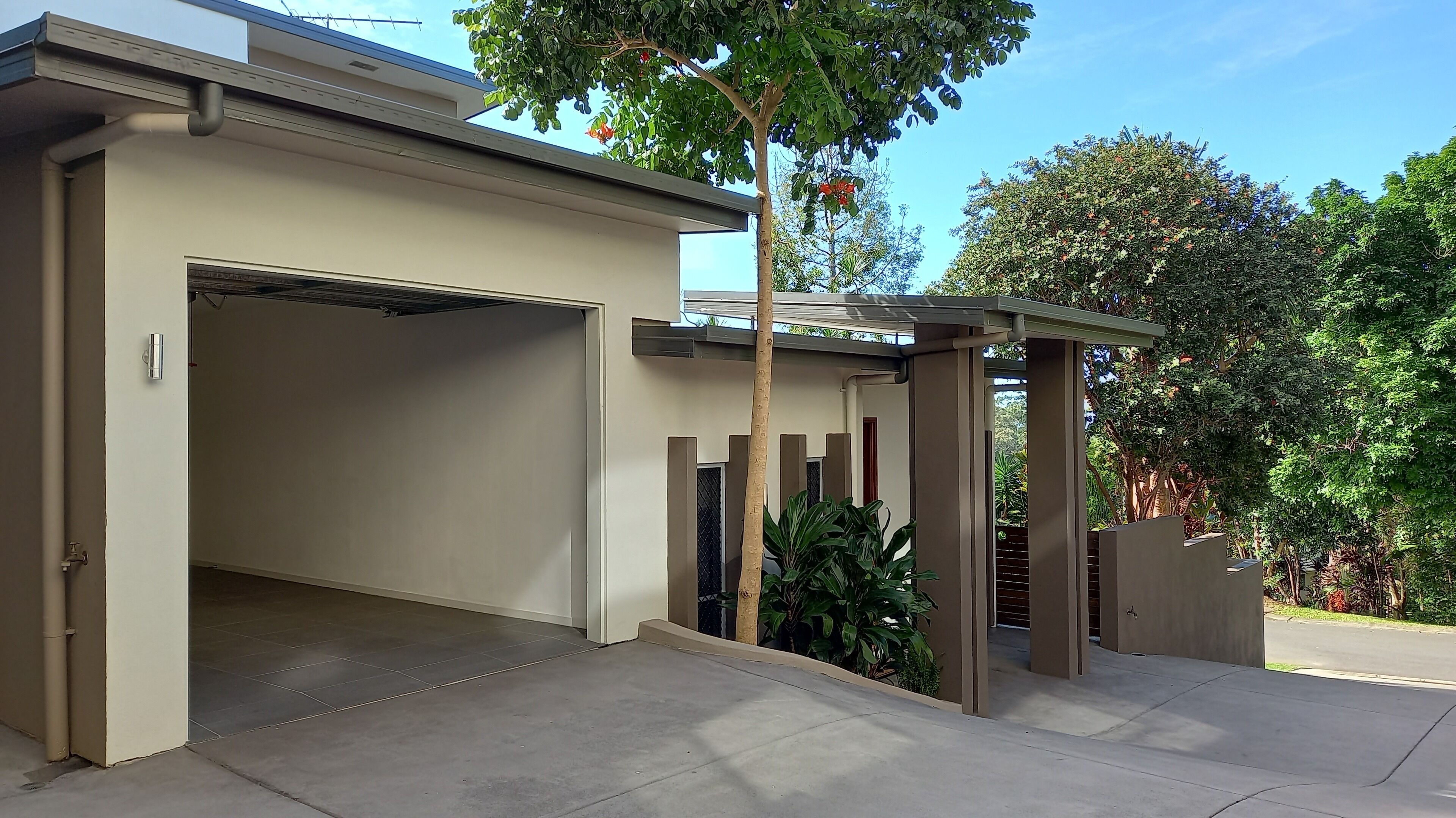 A modern garage with a carport, a concrete driveway, and a tree with green foliage.