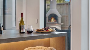 A modern kitchen with a wine bottle, glass, and fruit on the counter, a fireplace in the background, and a cozy seating area with fur-covered stools. - Central Queenstown Villa & Spa (Queenstown)