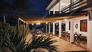 A modern outdoor patio area with wooden tables and chairs, surrounded by plants and a wooden pergola.