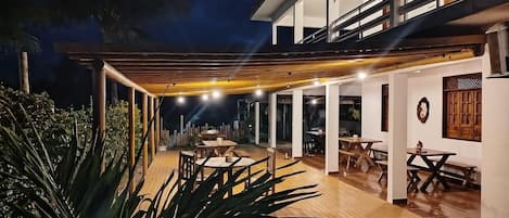 Un patio exterior moderno con mesas y sillas de madera, rodeado de plantas y una pérgola de madera.