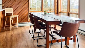 A room with a wooden sloped ceiling, a large dining table with chairs, and large windows with a view of trees.