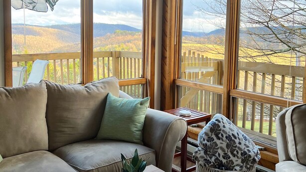A cozy room with a large window overlooking a wooden deck and hills. There's a sofa, a small table, and a patterned pouf.