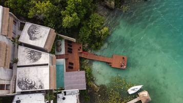 Vista aérea de una propiedad costera con una terraza de madera que se extiende sobre aguas turquesas cristalinas, rodeada de exuberante vegetación.