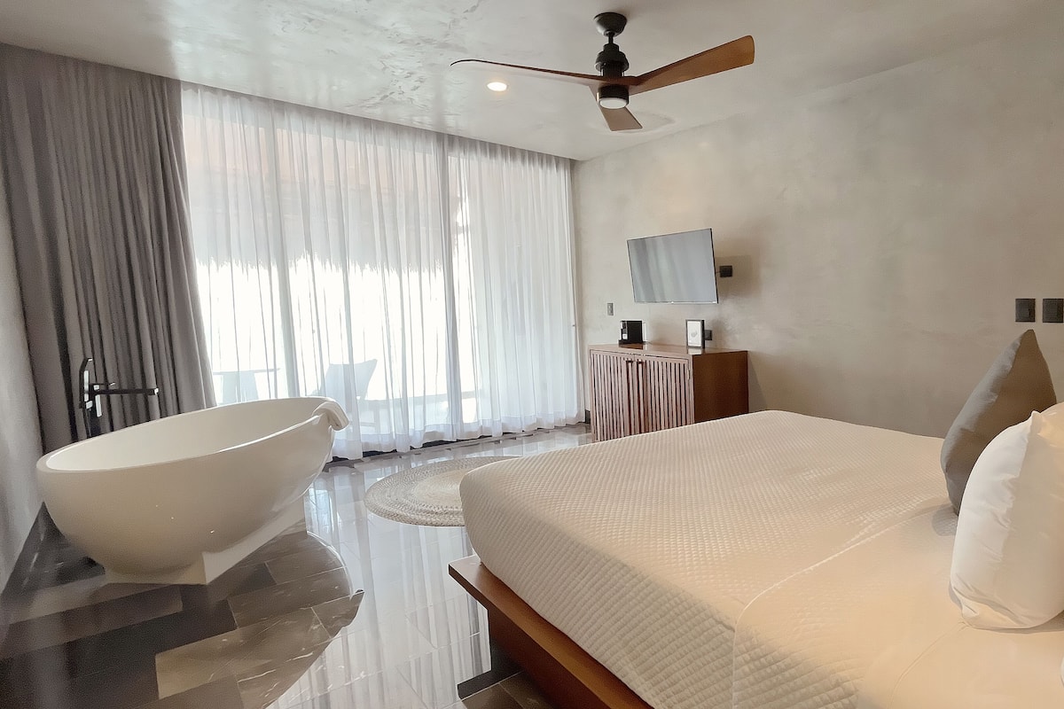 Un dormitorio moderno con una cama grande, una bañera independiente, un ventilador de techo y una alfombra redonda.