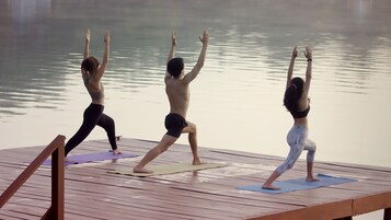 Drie personen beoefenen yoga op een houten terras aan een rustig waterlichaam.