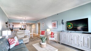A spacious living room with a grey sofa, a wooden coffee table, and a TV on a white cabinet. A dining area with white chairs and a kitchen in the background is visible.