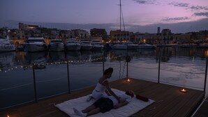 Een houten terras met een stel dat op een mat ligt, met uitzicht op een jachthaven met boten en gebouwen op de achtergrond.