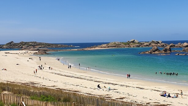 Una playa con aguas turquesas cristalinas, afloramientos rocosos y gente disfrutando de la arena y el agua.