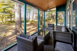 Un porche cubierto con mobiliario de mimbre, ventanas amplias y vistas a zona boscosa.