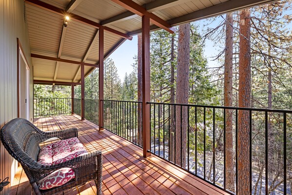A covered outdoor deck with a wicker chair and a table, surrounded by a metal railing, overlooking a forested area.
