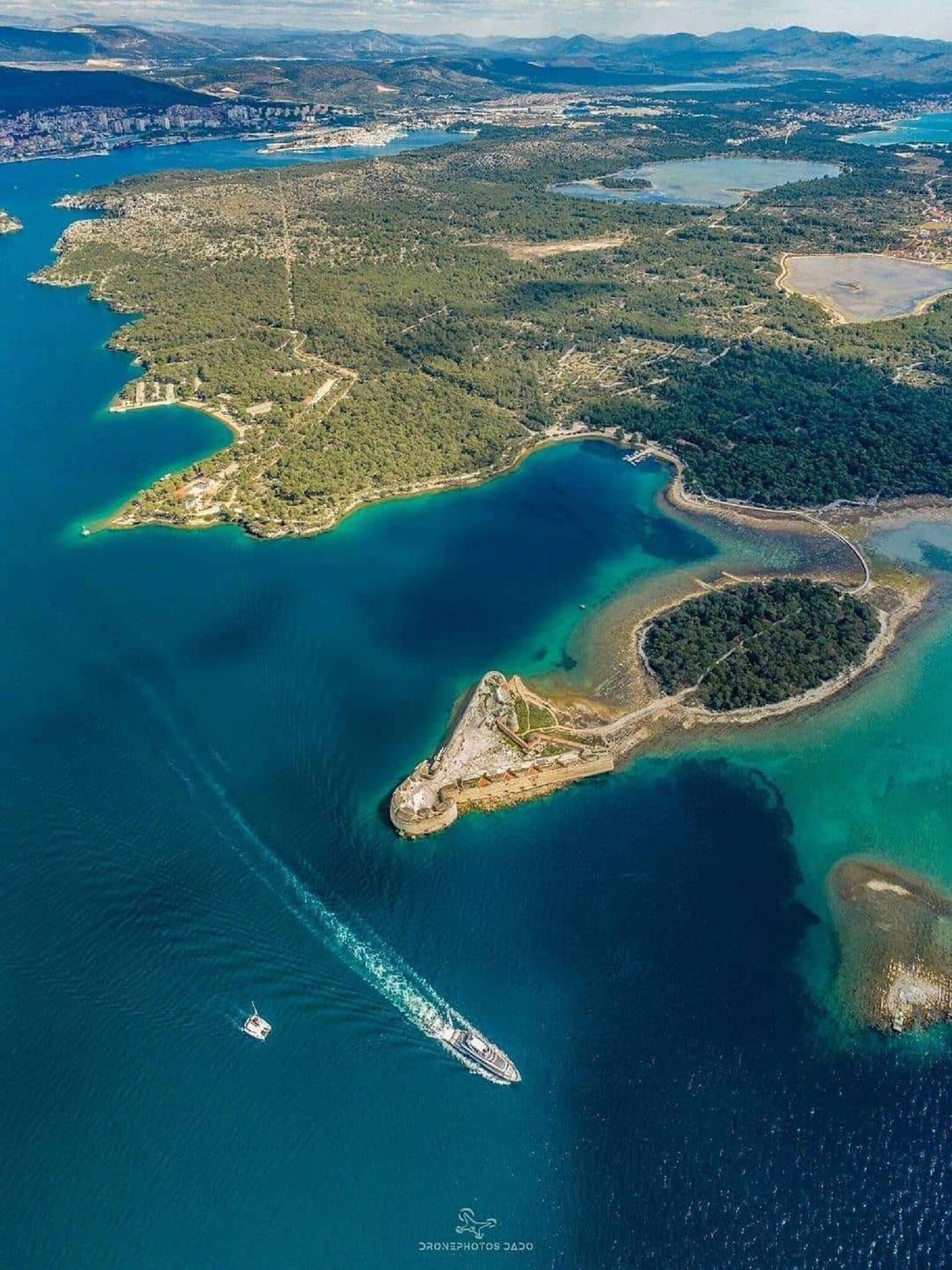 An aerial view of a coastal area with a marina, a small island with a fort, and surrounding greenery.
