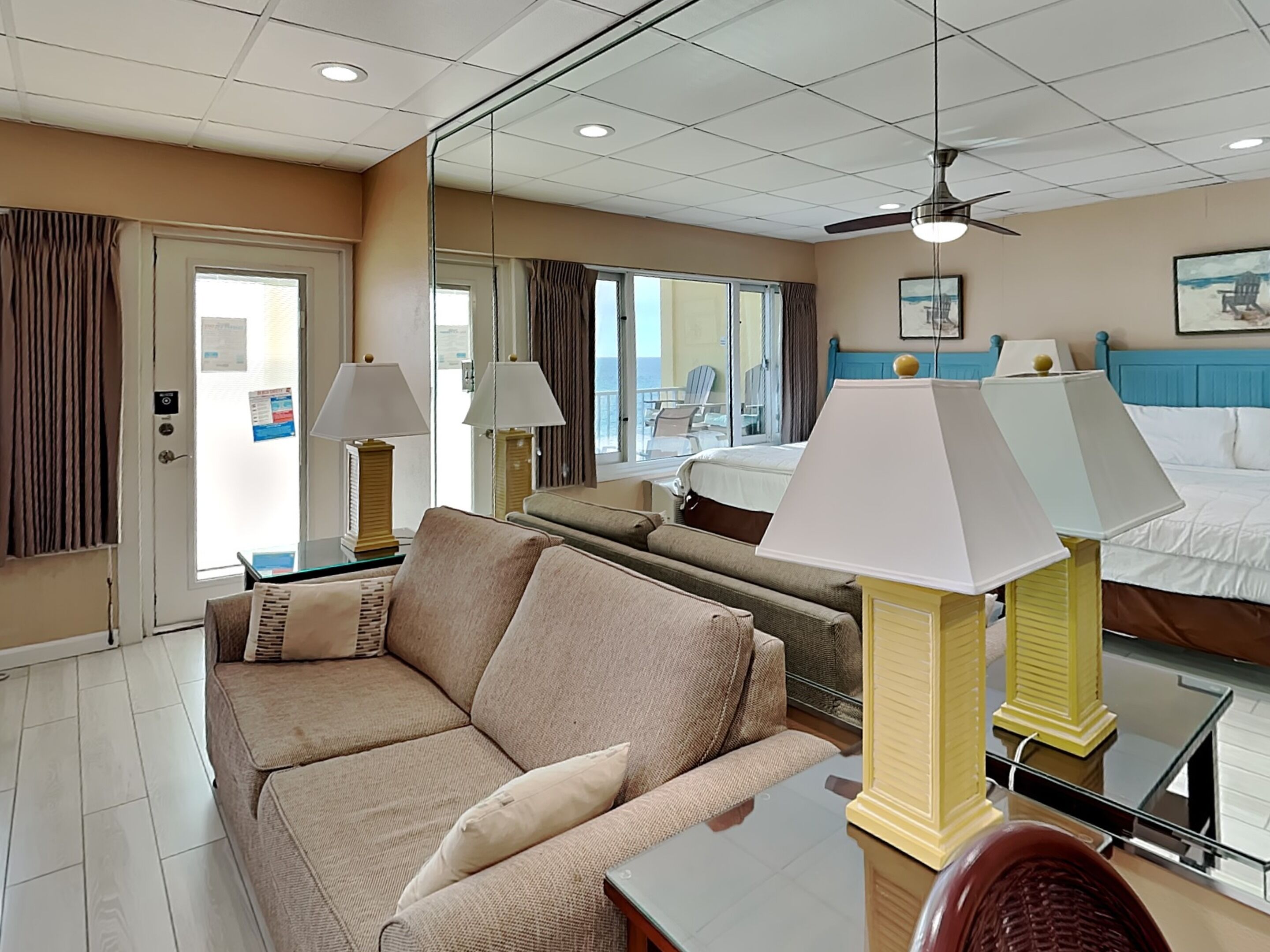 A hotel room with a sofa, two bedside tables with lamps, a bed, a ceiling fan, and a large window with curtains.