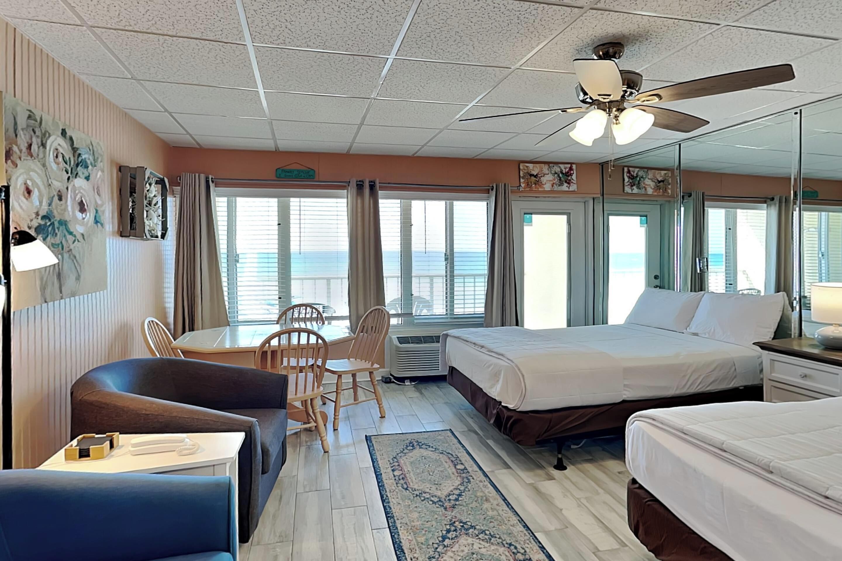 A hotel room with a bed, a desk with a chair, a sofa, a ceiling fan, and a view of the ocean.